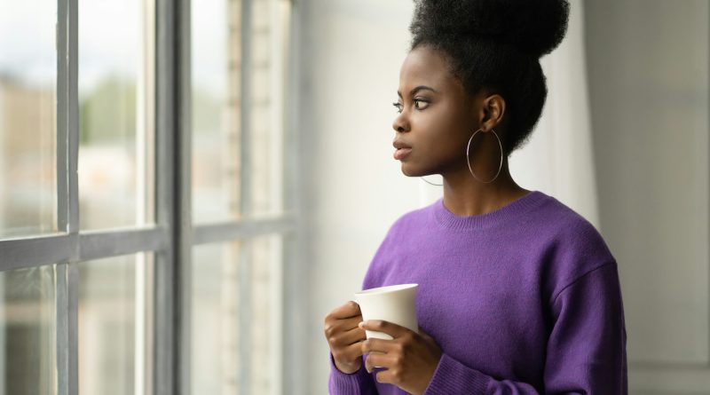 Pensive African American millennial woman thinking, looking through the window, holding mug coffee